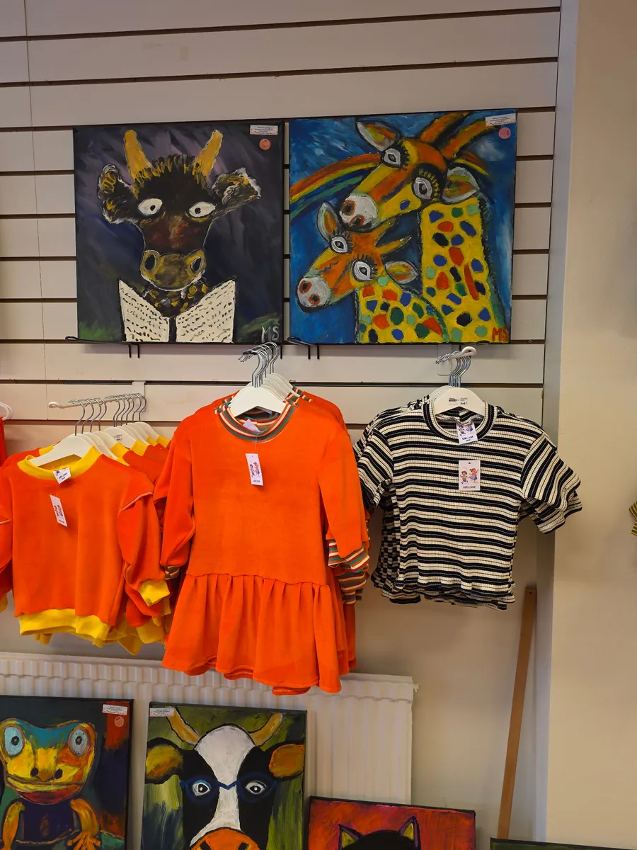Lekloop i butiken: orange sammetsklänning och tröja med gula muddar samt en randig tröja, hängande under två tavlor med en ko som läser och två giraffer