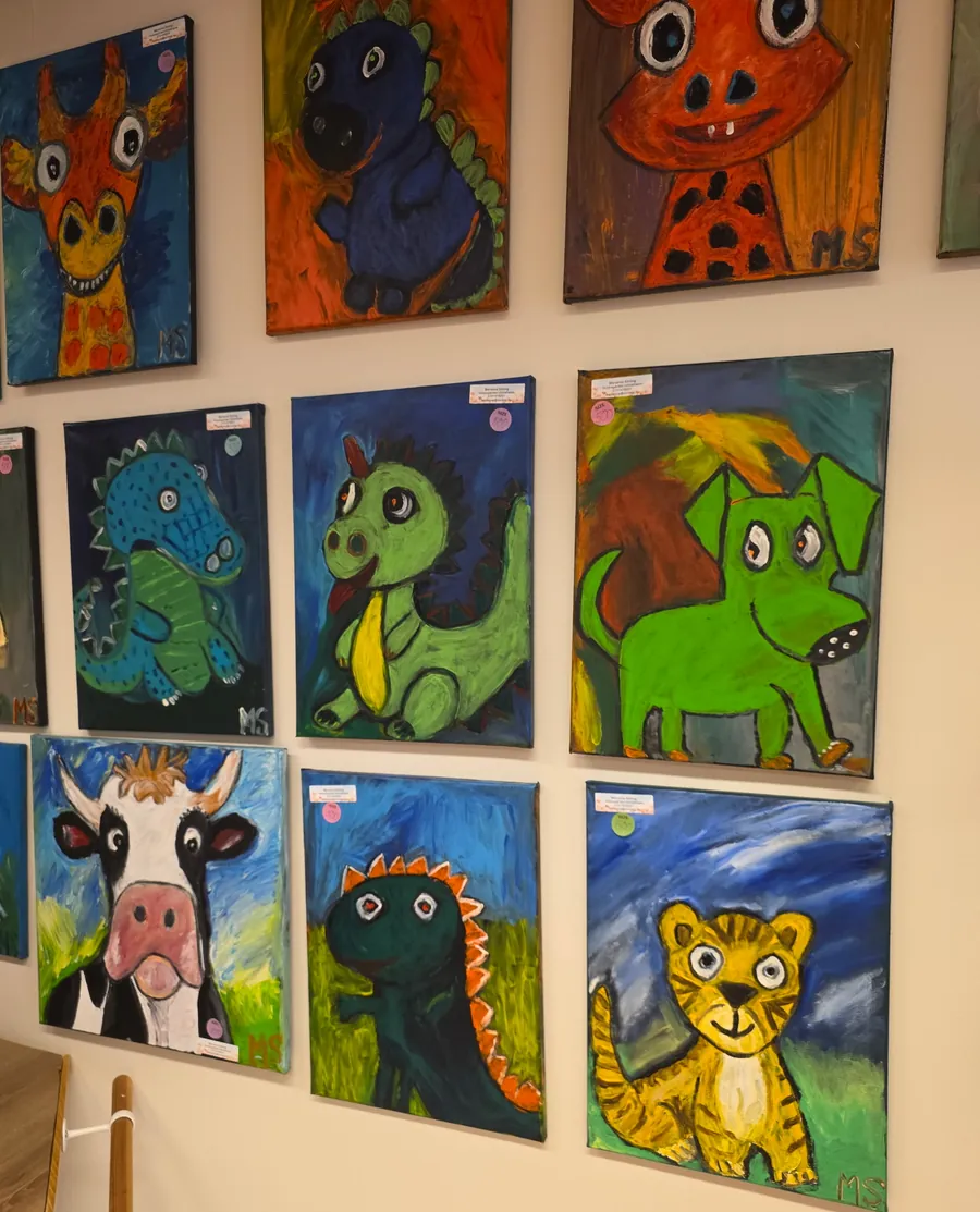 Vägg med tavlor på rad: en giraff, en blå dinosaurie, en grön dinosaurie med slips, en grön hund, en ko, en mörkgrön dinosaurie och en tiger